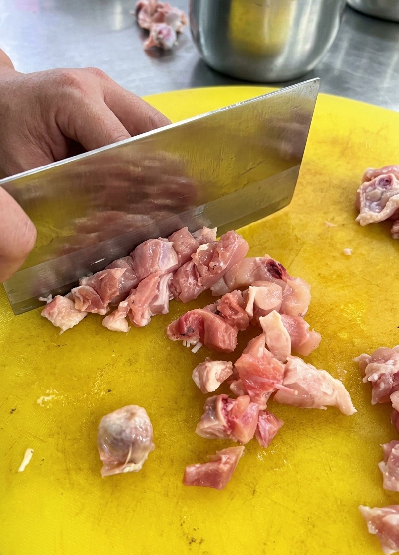 Eine Person schneidet rohes Hähnchenfleisch mit einem Beil in kleine Würfel auf einem gelben Kunststoffbrett.