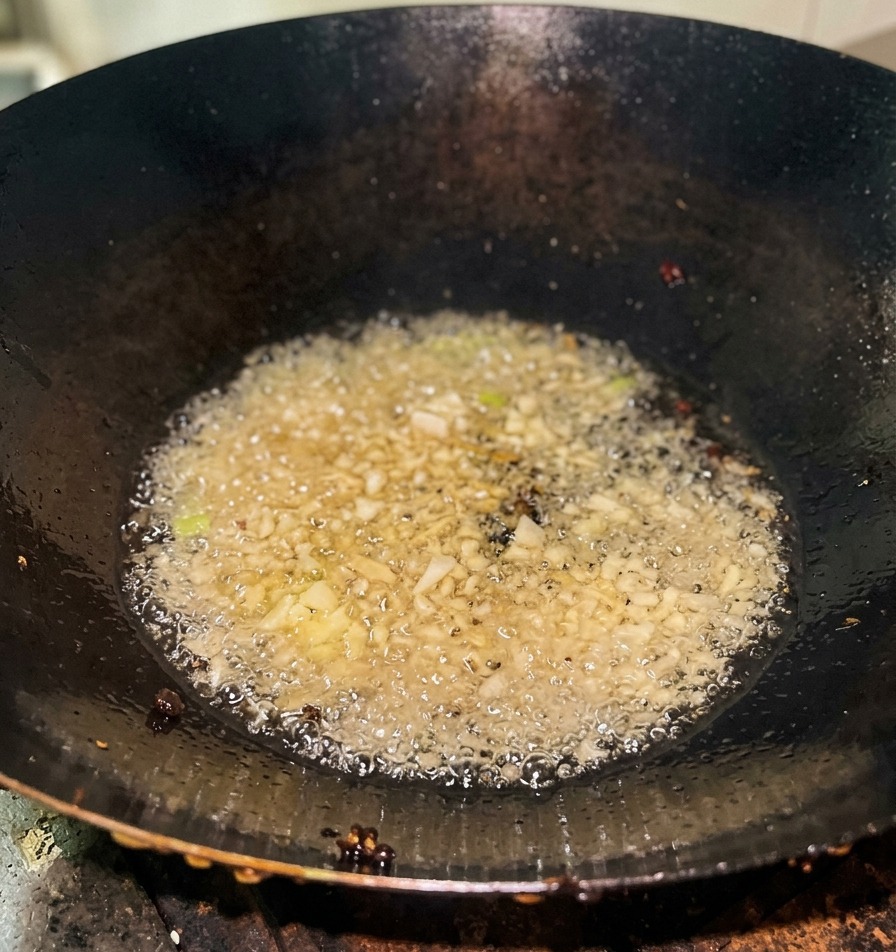Gehackte Frühlingszwiebeln brutzeln in klarem heißem Öl am Boden eines dunklen Eisen-Woks.