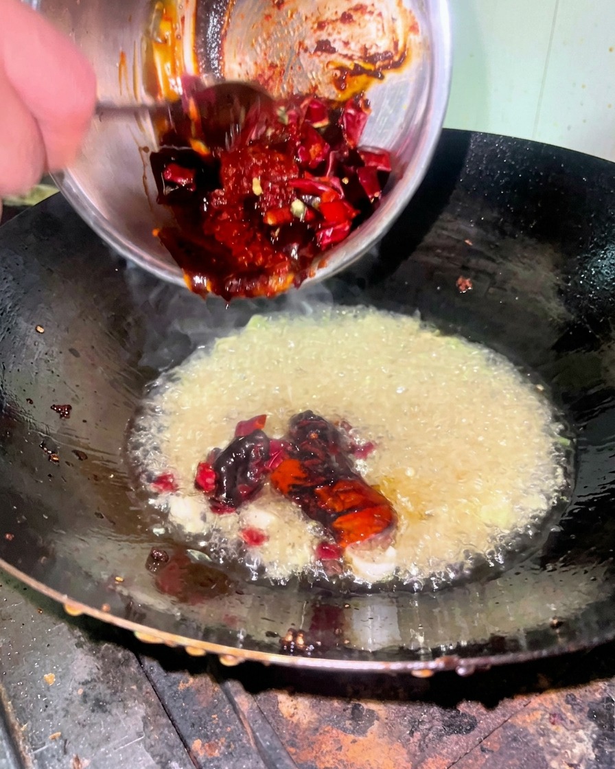 Eine dicke, dunkelrote Saucenmischung wird aus einer Metallschüssel in einen Wok mit heißem Öl gegeben.