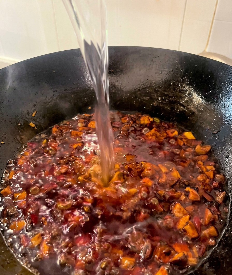 Ein Wasserstrahl wird in einen Wok mit dunkelroter, köchelnder Hähnchen-Pilz-Sauce gegossen.