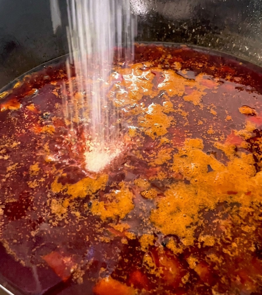 Weißes Würzpulver wird in eine tiefrote, sprudelnde Brühe mit Pilzen und Hähnchen gestreut.