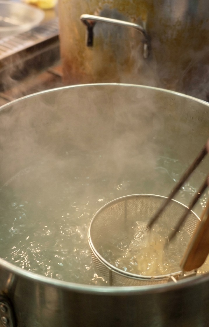 Ein Koch taucht weiße Ramen-Nudeln in einem Sieb in einen Topf mit kochendem Wasser.