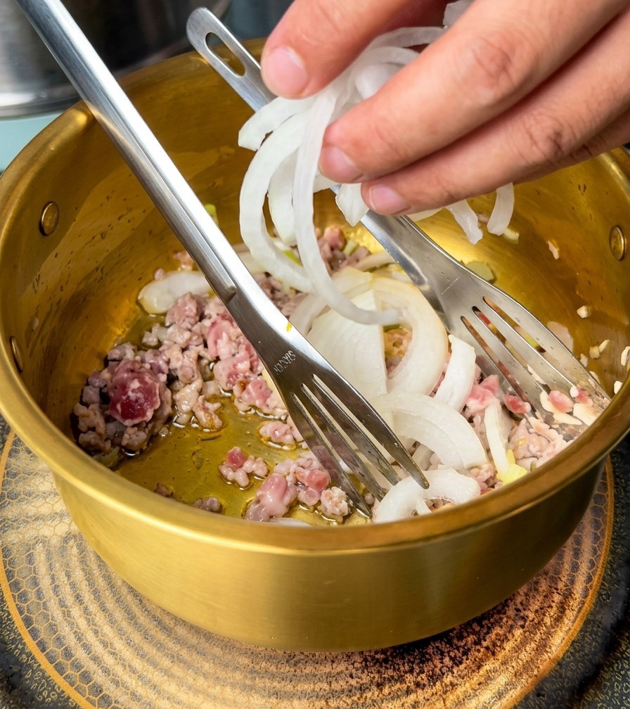 Eine Hand gibt weiße Zwiebelringe zu angebratenem Hackfleisch und Aromaten in den Topf.