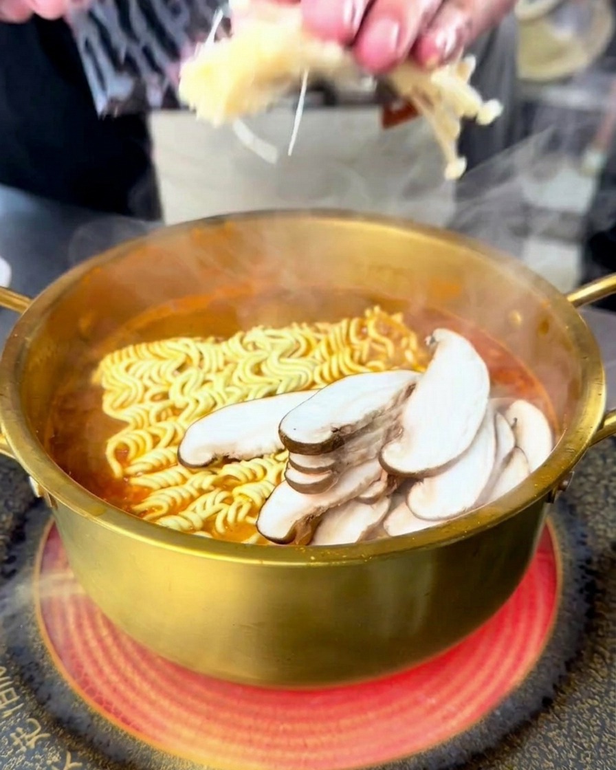 Eine Person gibt Enoki- und Shiitake-Pilze in den kochenden Topf mit Seafood-Ramen.