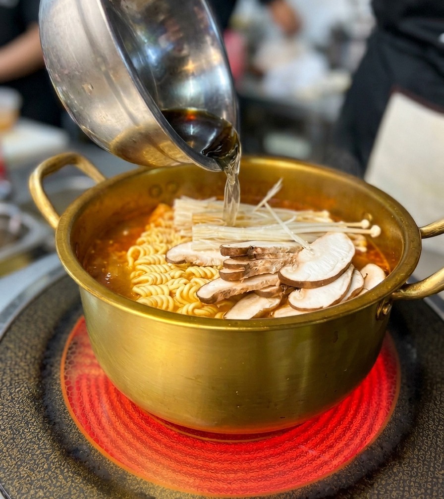 Eine Schale mit Pilzen und Meeresfrüchten wird in den goldenen Topf mit Nudeln und roter Brühe gegeben.