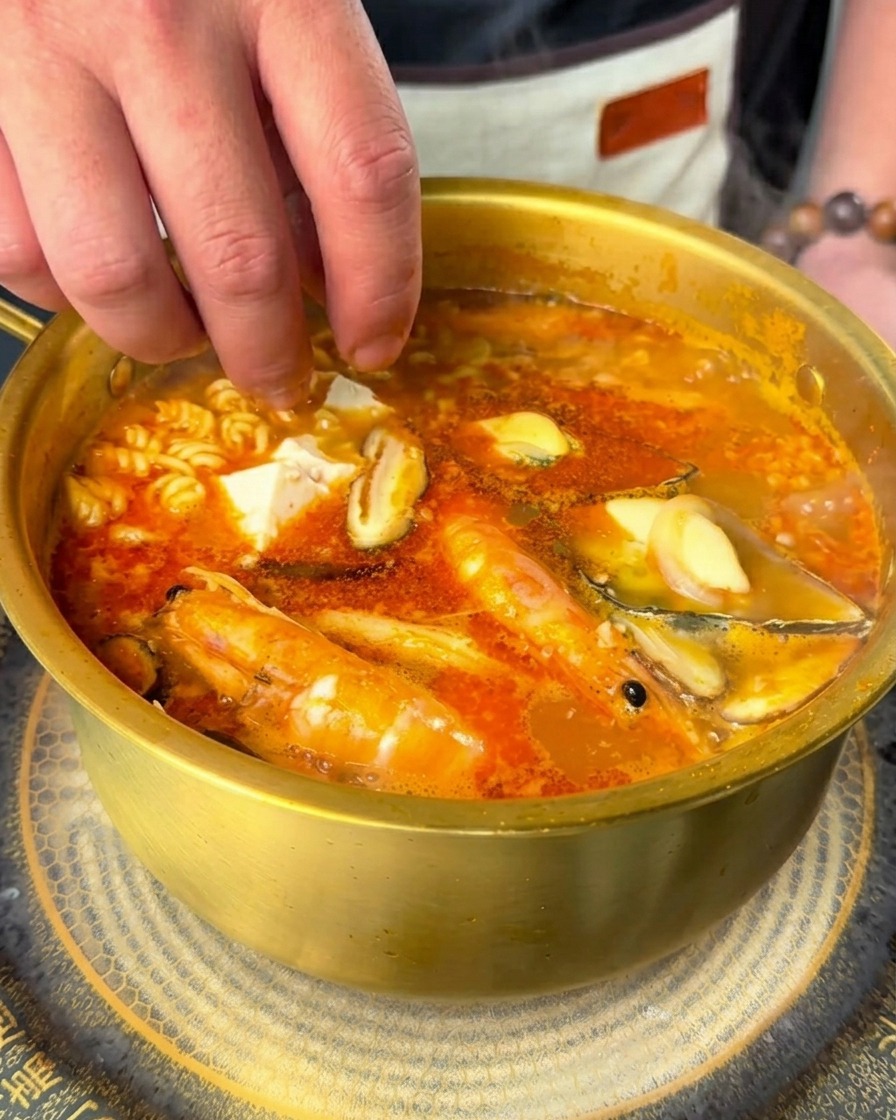 Eine Hand gibt weiße Seidentofuwürfel und frische Chiliringe in die sprudelnde Suppe.