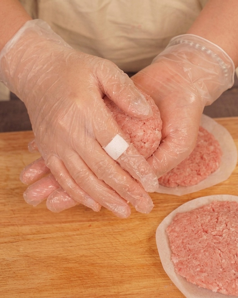 Eine Person mit Kunststoffhandschuhen wirft einen Ball aus rohem Hackfleisch über einem Holzbrett zwischen den Händen hin und her.