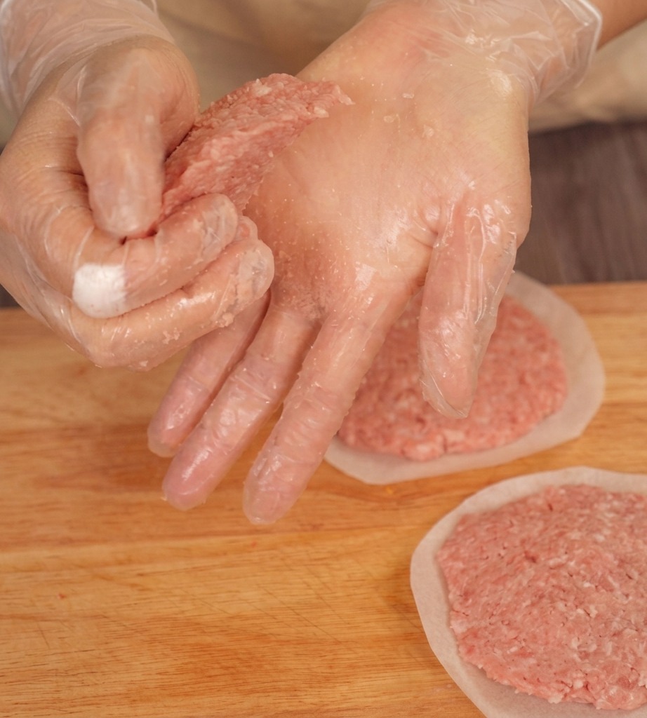 Hände drücken die Ränder eines rohen Beef-Patties auf einem Brett flach, um eine konische Form zu erzeugen.