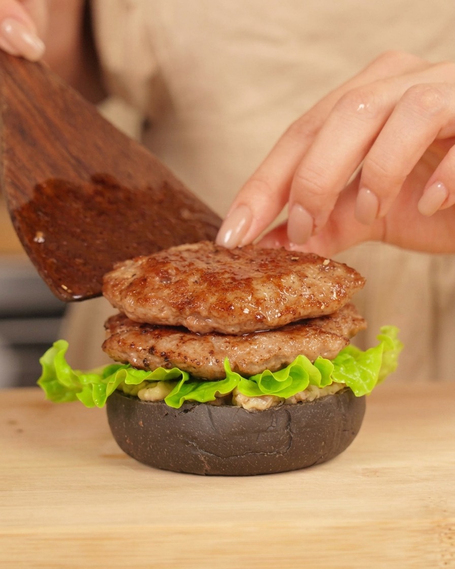 Eine Hand legt mit einem Pfannenwender vorsichtig ein zweites gebratenes Patty auf einen halb fertigen Burger.