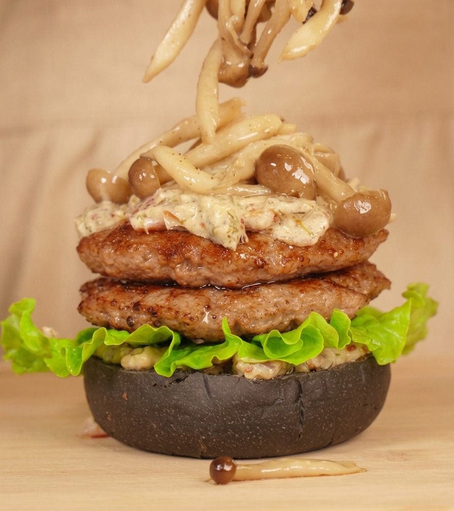 Eine Küchenzange platziert vorsichtig Buchenpilze auf dem Burgerstapel, der mit cremiger Sauce überzogen ist.