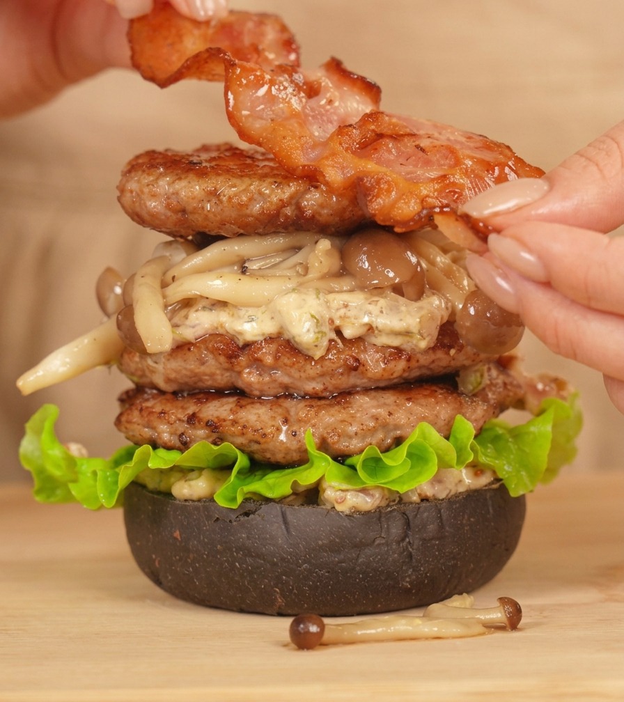 Hände legen einen Streifen perfekt gebratenen Speck auf einen hohen, fertig zusammengesetzten Double-Beef-Burger.