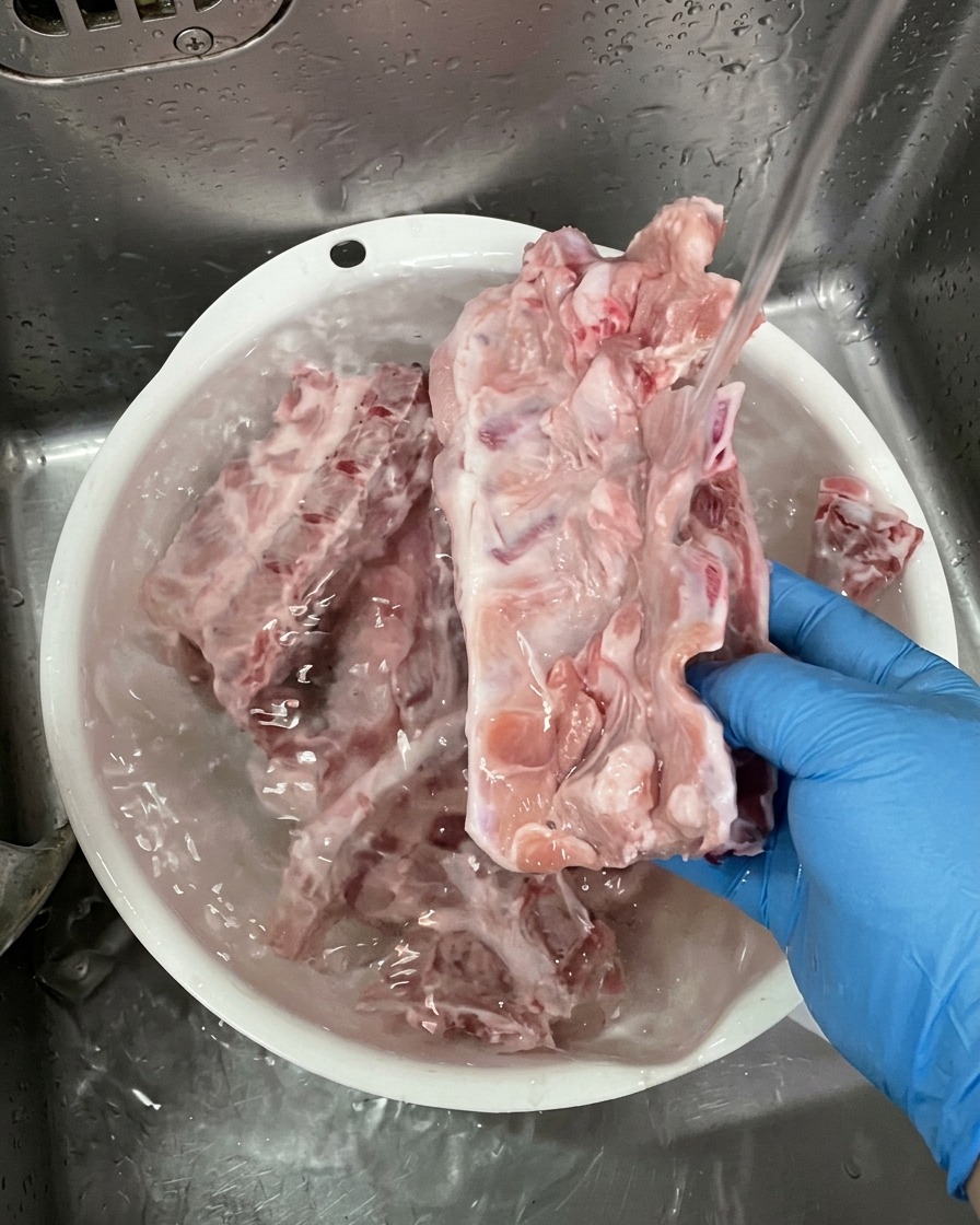 Person mit blauen Handschuhen wäscht rohe Schweineknochen unter fließendem Wasser in einer weißen Schüssel.