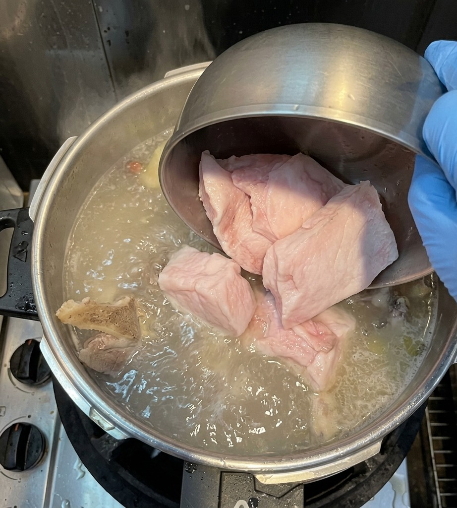 Eine Hand gibt große Stücke weißes Schweinerückenfett in den kochenden Topf.