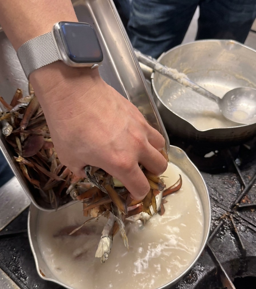 Die Hand eines Kochs gibt getrocknete Fischzutaten in einen Topf mit weißer Brühe.