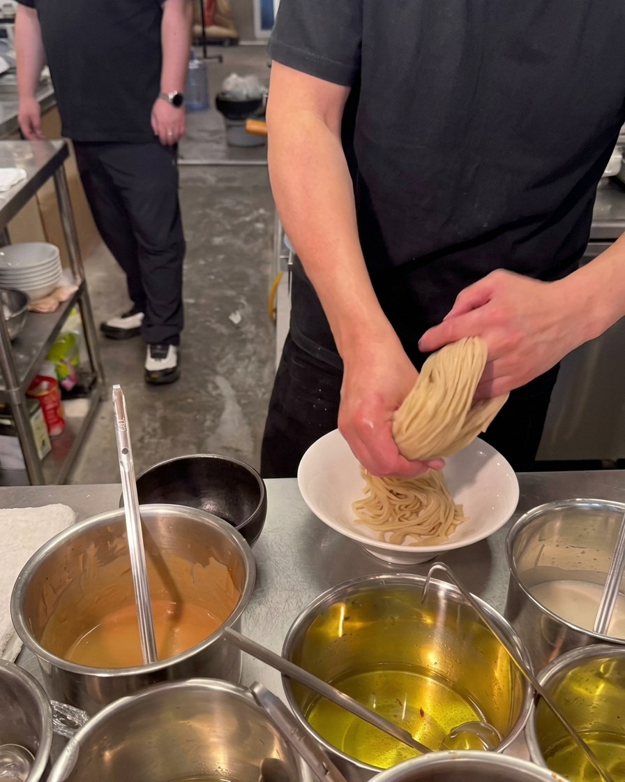 Ein Koch faltet dicke Ramen-Nudeln mit beiden Händen in eine weiße Keramikschüssel.