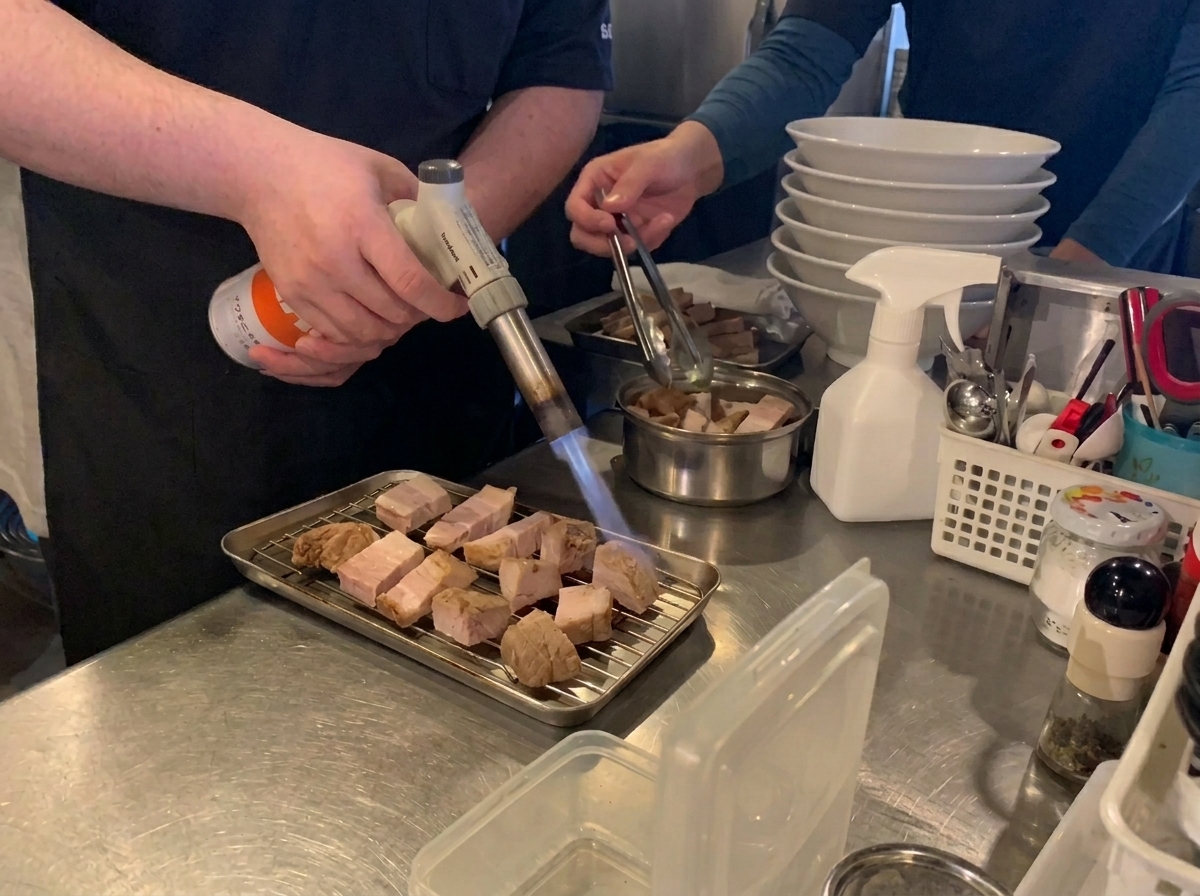 Ein Koch nutzt einen Küchenbrenner, um Scheiben von Schweinebauch auf einem Rost zu karamellisieren.