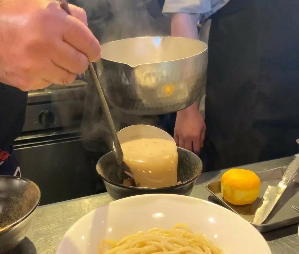 Dicke, cremige Ramen-Brühe wird aus einer Metallkelle in eine schwarze Keramikschale gegossen.