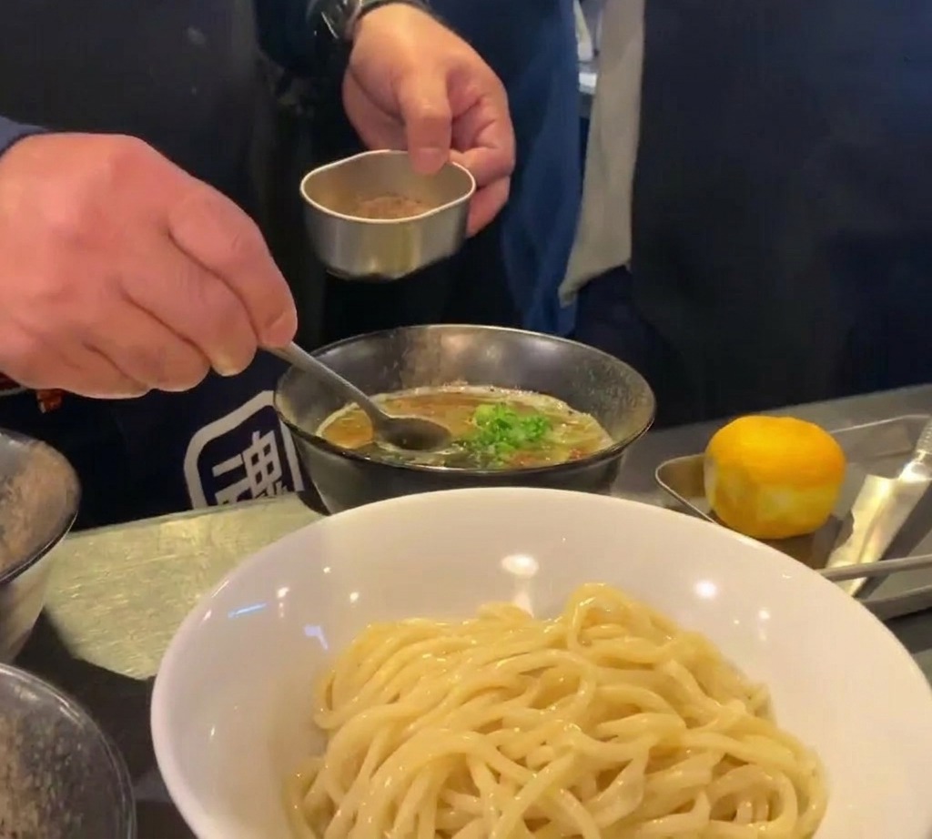 Ein Koch streut feines braunes Fischpulver über eine Schale mit dunkler Dip-Suppe.