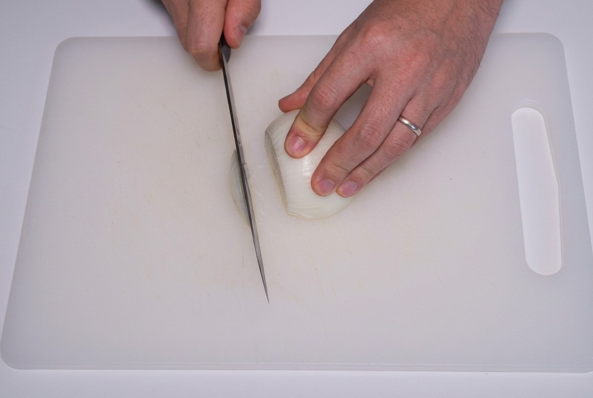 Hände, die eine weiße Zwiebel mit einem scharfen Messer in dünne Streifen auf einem Schneidebrett schneiden.