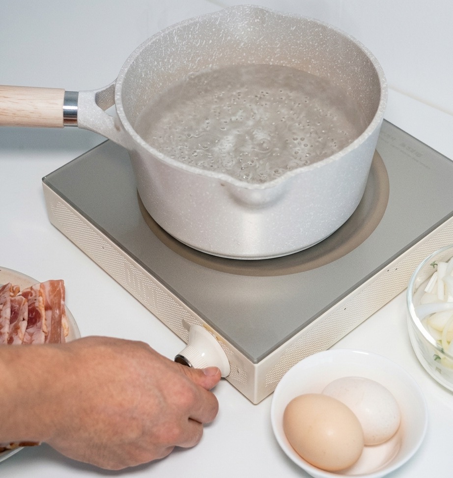 Eine Hand, die den Regler einer Induktionsplatte dreht, auf der ein Topf mit Wasser steht.