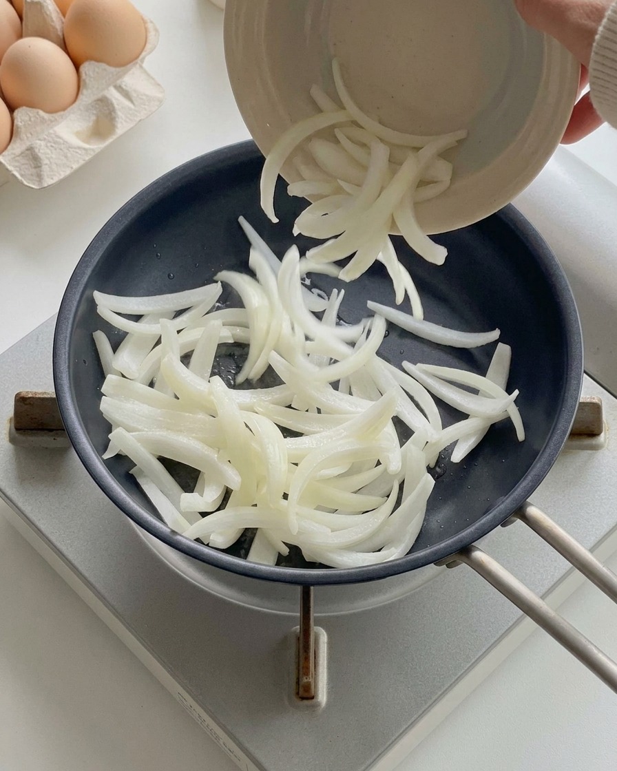 Geschnittene weiße Zwiebeln werden aus einer kleinen Schüssel in eine erhitzte schwarze Pfanne geschüttet.