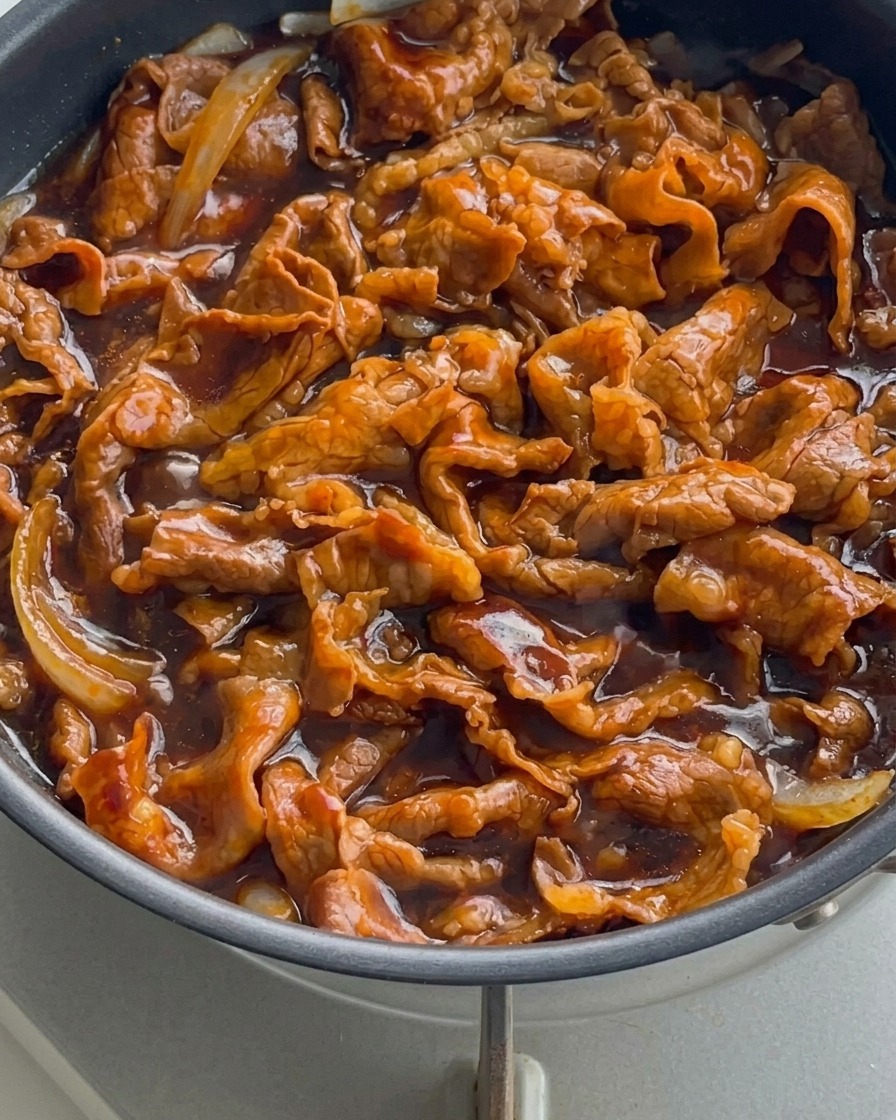 Nahaufnahme von Rindfleisch und Zwiebeln, die in einer dicken, glänzenden Sauce in der Pfanne brutzeln.