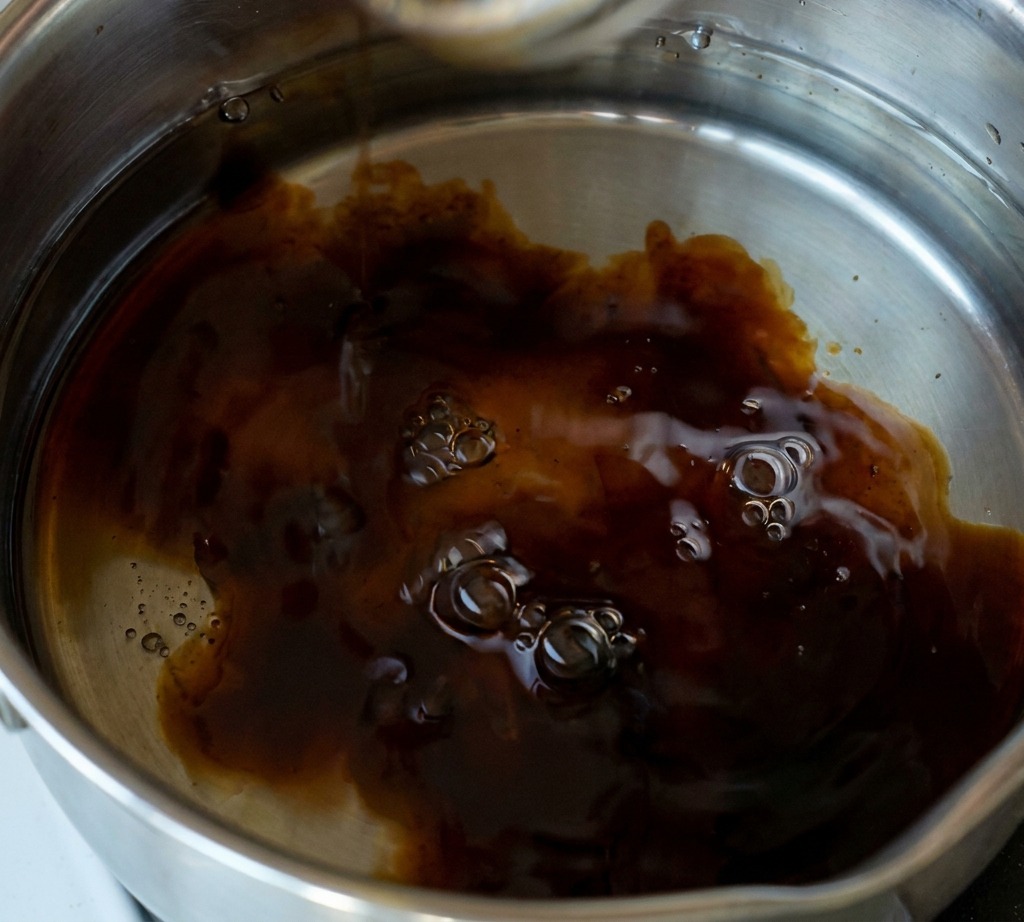 Eine Hand gibt einen Löffel weißen Zucker in einen Edelstahltopf mit einer dunklen, köchelnden Sauce auf Sojabasis.