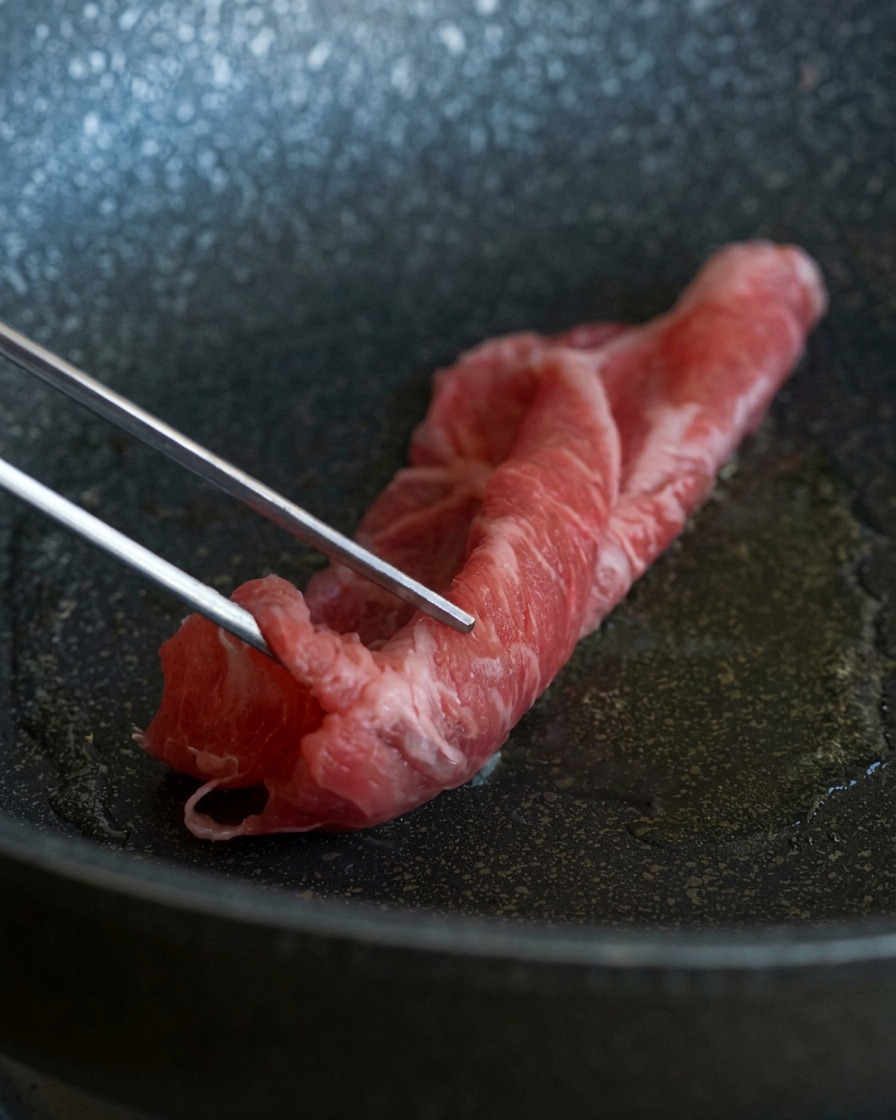 Mit einer Metallzange wird eine dünne, marmorierte Scheibe rohes Rindfleisch in eine heiße Pfanne mit schimmerndem Öl gelegt.