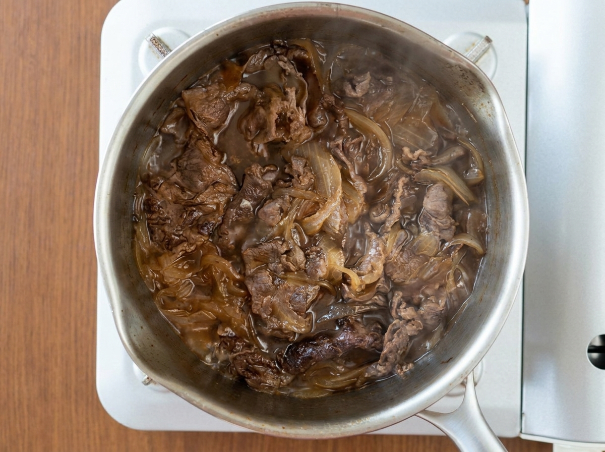 Draufsicht auf Rindfleischscheiben und Zwiebeln, die zusammen in einer dunklen, blubbernden Sauce in einem Edelstahltopf köcheln.