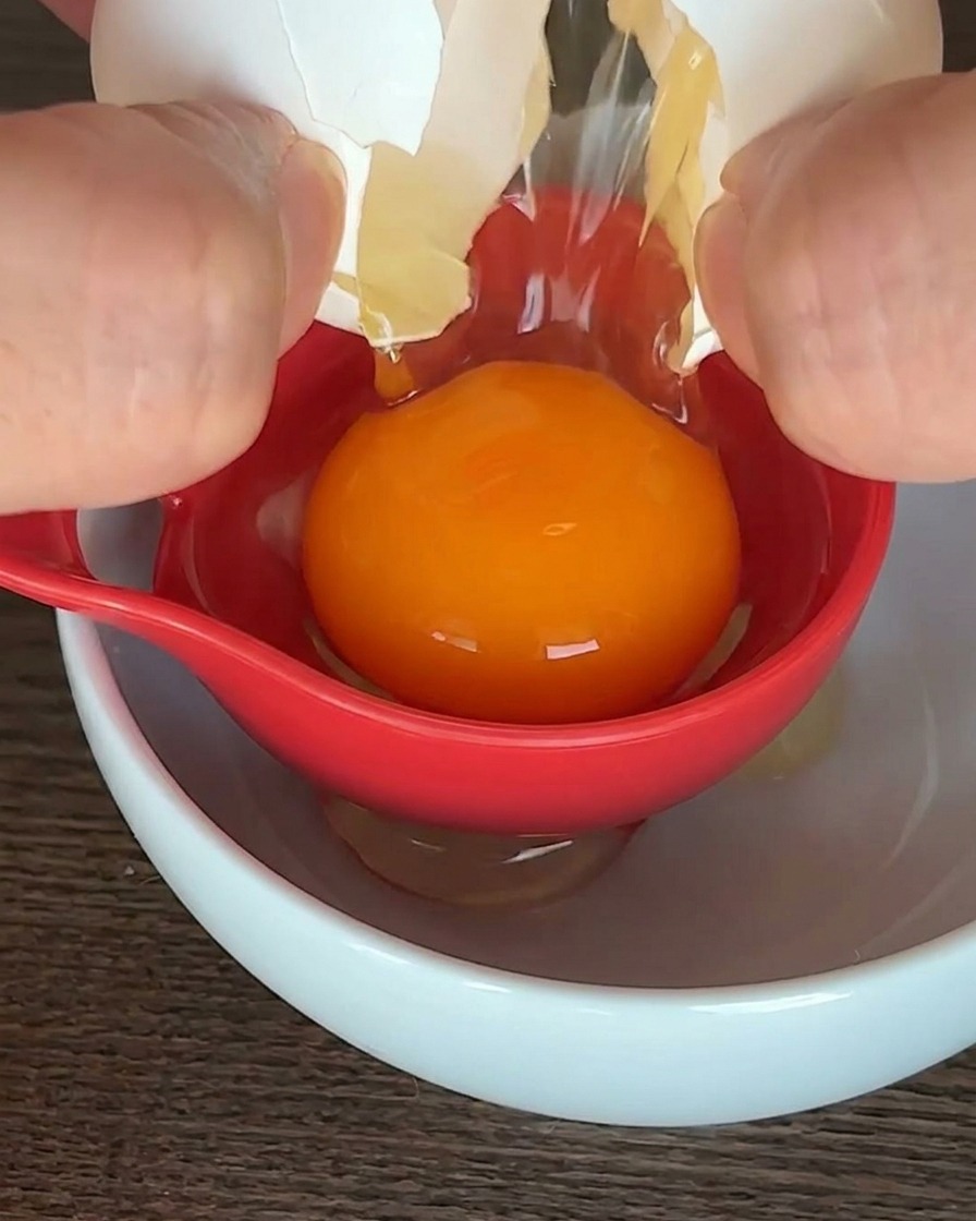 Ein rohes Ei wird über einer weißen Schüssel mit einem roten Eitrenner getrennt, das orangefarbene Eigelb bleibt in der Mitte liegen.