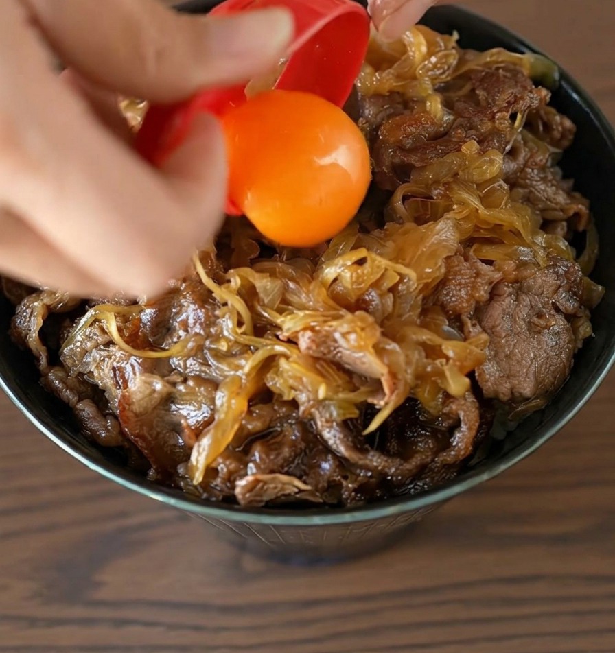Ein leuchtend orangefarbenes Eigelb gleitet aus einem roten Eitrenner direkt in die Mitte der fertigen Rindfleisch-Bowl.