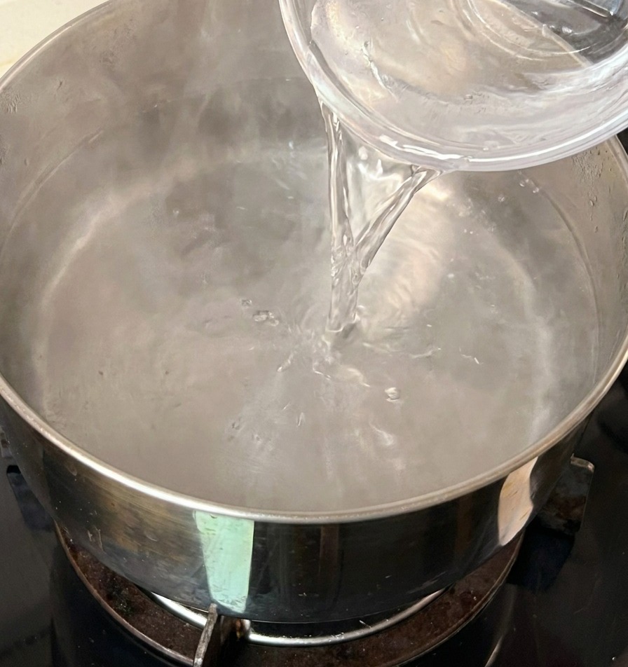 Kaltes Wasser wird aus einer kleinen Glasschüssel in einen Edelstahltopf mit heißem Wasser gegossen.
