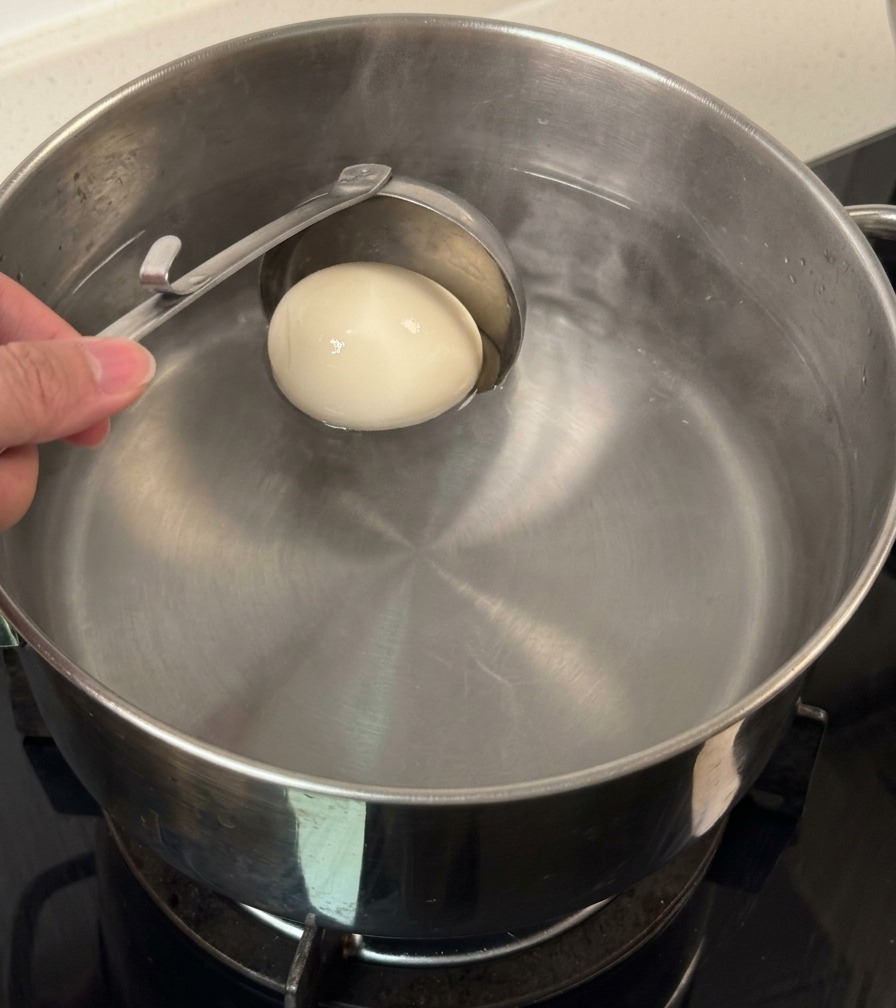 Ein einzelnes weißes Ei wird mit einer Metallkelle in einen Wassertopf gelassen.
