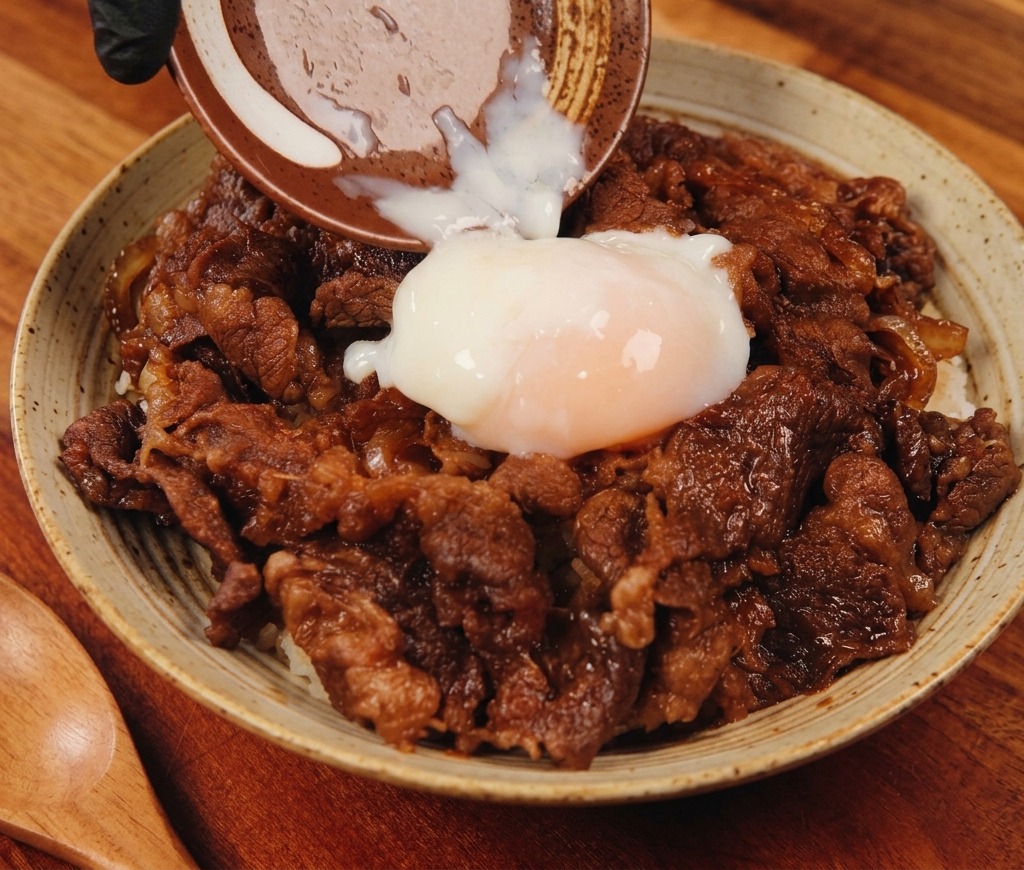 Ein weichgekochtes Onsen-Ei wird aus einer kleinen Keramikschale auf eine dampfende Bowl mit herzhaftem Rindfleisch und Reis gleiten gelassen.