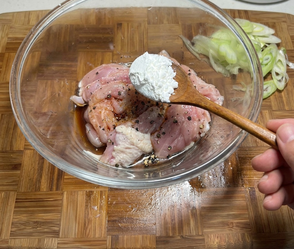 Ein Löffel Speisestärke wird zu einer Schüssel mit rohem Hähnchenfleisch gegeben, das in Sojasauce und Gewürzen mariniert ist.