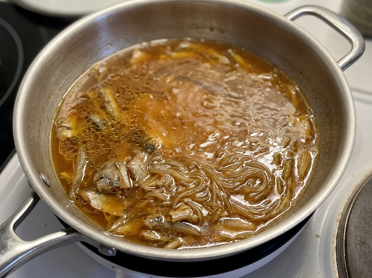 Ein Glasdeckel wird auf eine Pfanne gesetzt, in der Hähnchen und Zwiebeln in einer dunklen Sauce schmoren.