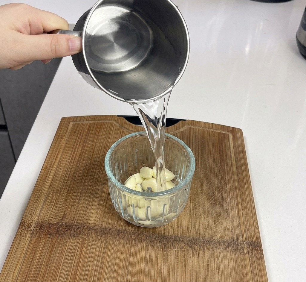 Ein Metallbecher gießt Wasser in einen Mixer voller frischer, weißer Knoblauchzehen.