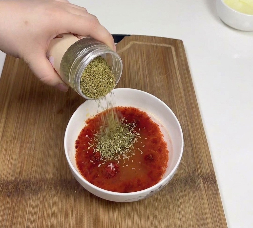 Eine Hand gießt gemahlenen Kreuzkümmel aus einem Glas in eine Schüssel mit der roten Chilisaucen-Basis.