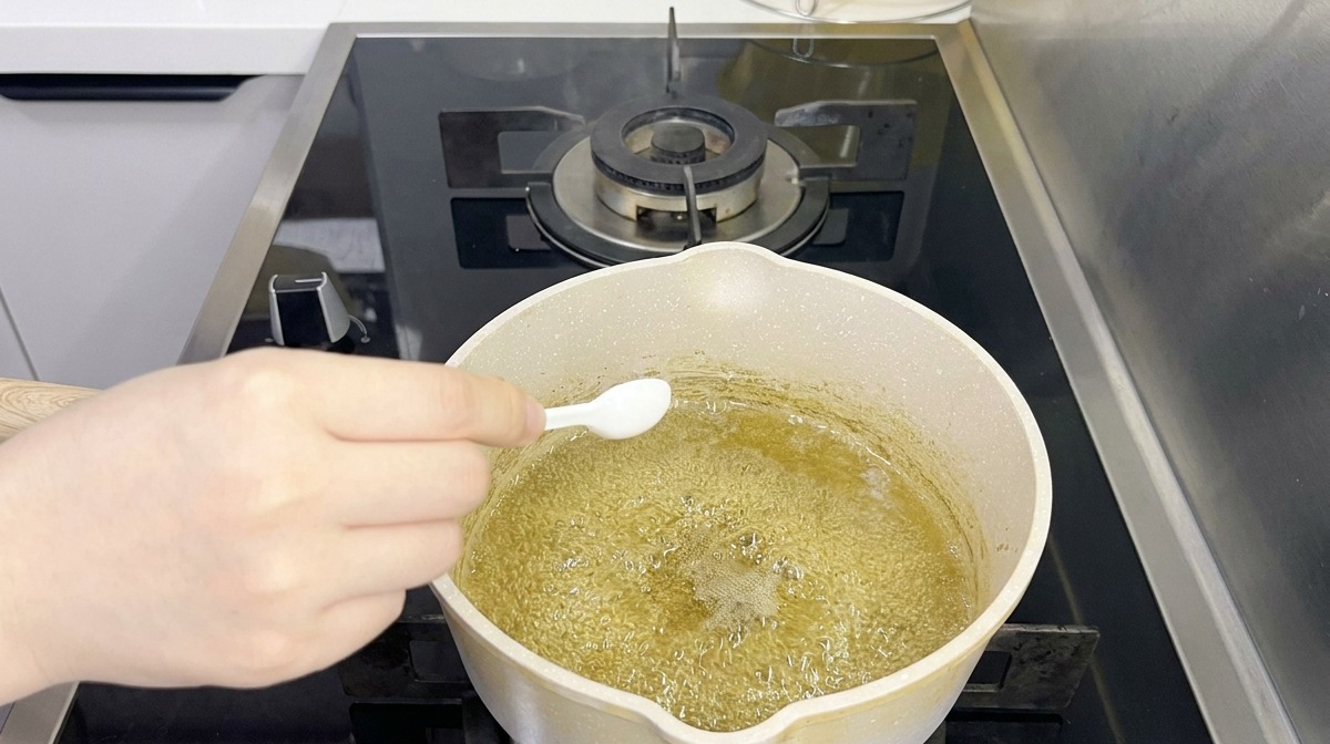 Eine Person gibt mit einem kleinen weißen Löffel Gewürze in eine kochende, grünliche Flüssigkeit in einem Topf.