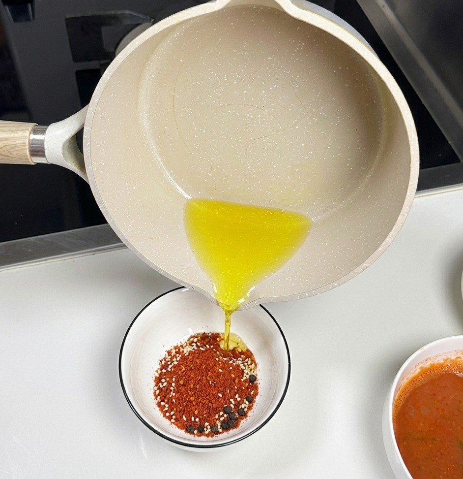 Ein heller Keramiktopf gießt klares heißes Öl in eine kleine weiße Schüssel mit rotem Chilipulver und weißem Sesam.