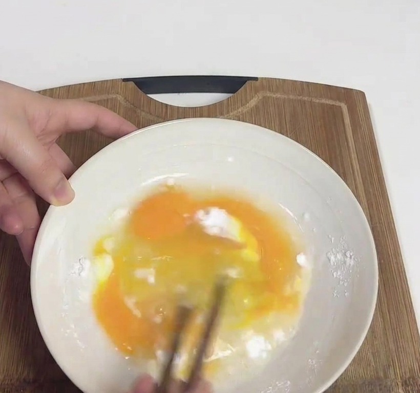 Hände verquirlen Eier und weiße Kartoffelstärke mit Holzstäbchen in einer weißen Schüssel.