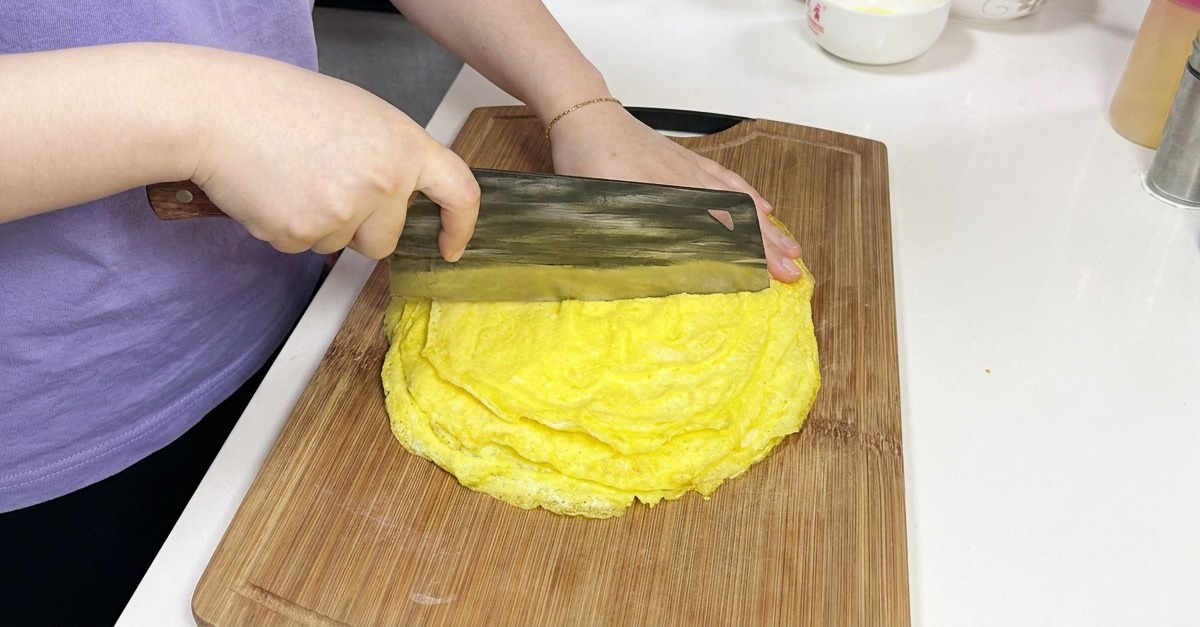 Eine Person schneidet mit einem großen chinesischen Hackmesser einen Stapel dünner Eierpfannkuchen auf einem Holzbrett.