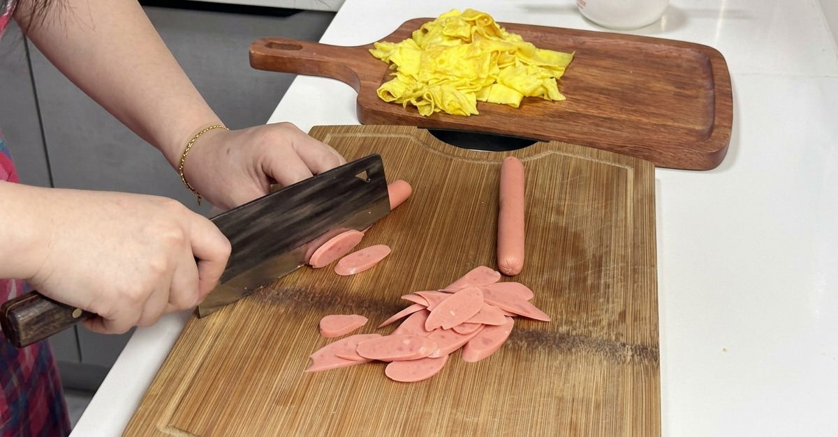 Eine Person schneidet rosa Würstchen schräg mit einem Hackmesser auf einem Holzschneidebrett.