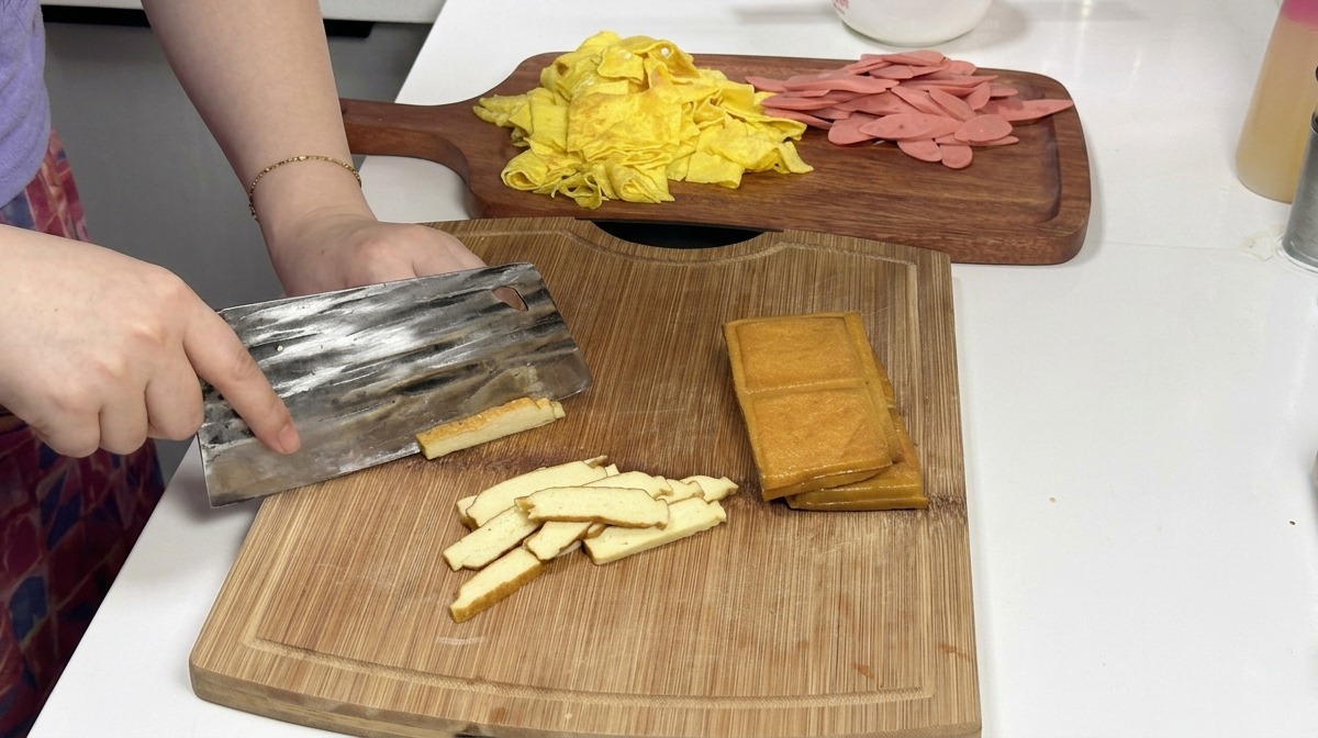 Ein Metallhackmesser schneidet durch einen Stapel goldbrauner Räuchertofublöcke auf einem Holzbrett, im Hintergrund liegen vorbereitete Eier- und Schinkenstreifen.