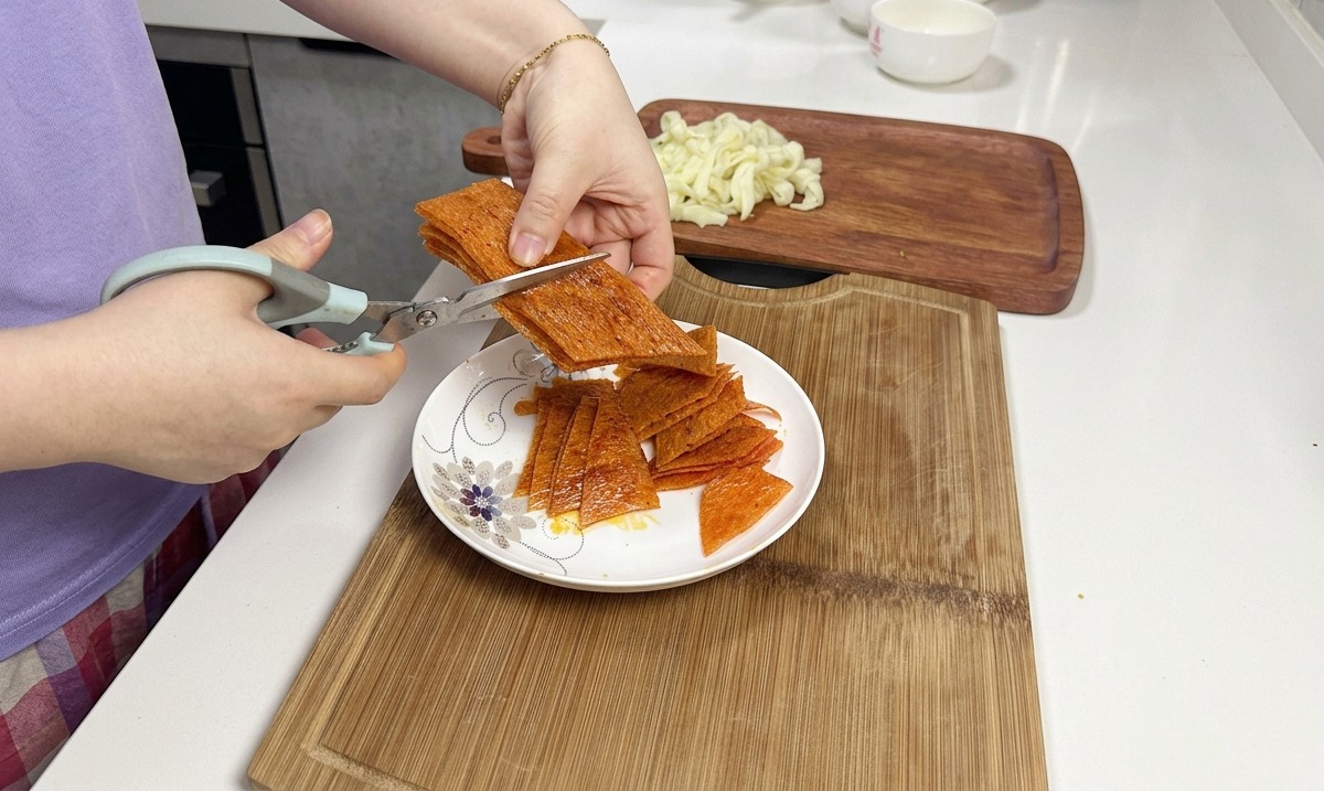 Eine Küchenschere schneidet einen Stapel orangefarbener, scharfer Latiao-Streifen über einem weißen Keramikteller.