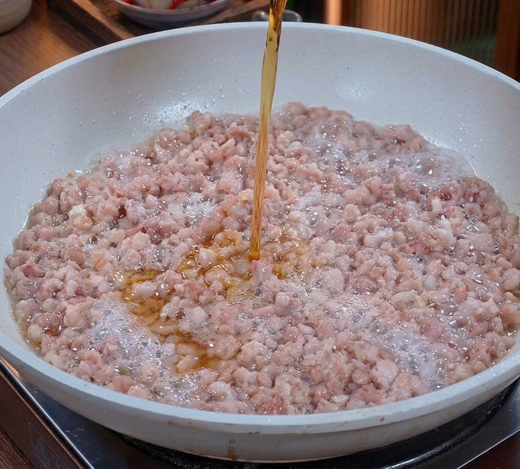 Eine klare, bernsteinfarbene Flüssigkeit wird aus einem Glas über das bratende Hackfleisch in der Pfanne gegossen.