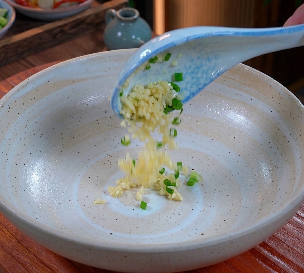 Gehackter Knoblauch und Frühlingszwiebeln fallen von einem blau-weißen Löffel auf den Boden einer großen Keramikschüssel.