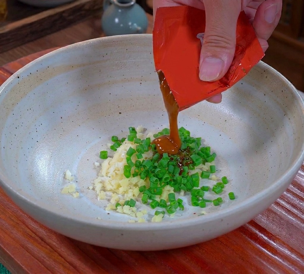 Eine Hand drückt dicke, braune Dandan-Sauce aus einer roten Verpackung in eine Keramikschüssel über gehackten Knoblauch und Frühlingszwiebeln.