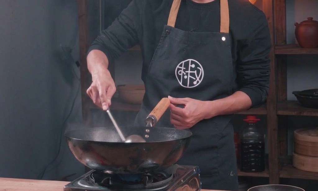 Ein Koch brät Hackfleisch in einem traditionellen schwarzen Wok an und gibt flüssige Würze hinzu.