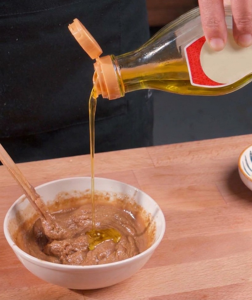 Klares goldenes Öl wird aus einer Flasche in eine weiße Schüssel mit dicker brauner Sesamsauce gegossen und mit einem Stäbchen verrührt.